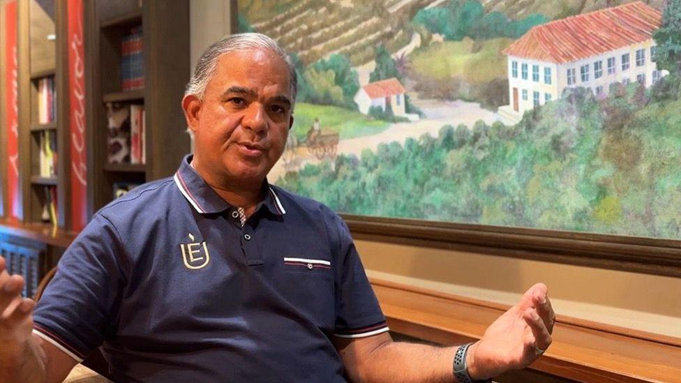 A seated half length portrait of Luiz. His arms are outstretched, he is wearing a dark blue polo shirt. Behind him on the wall is a large painting showing a rural scene with buildings, hills and fields.