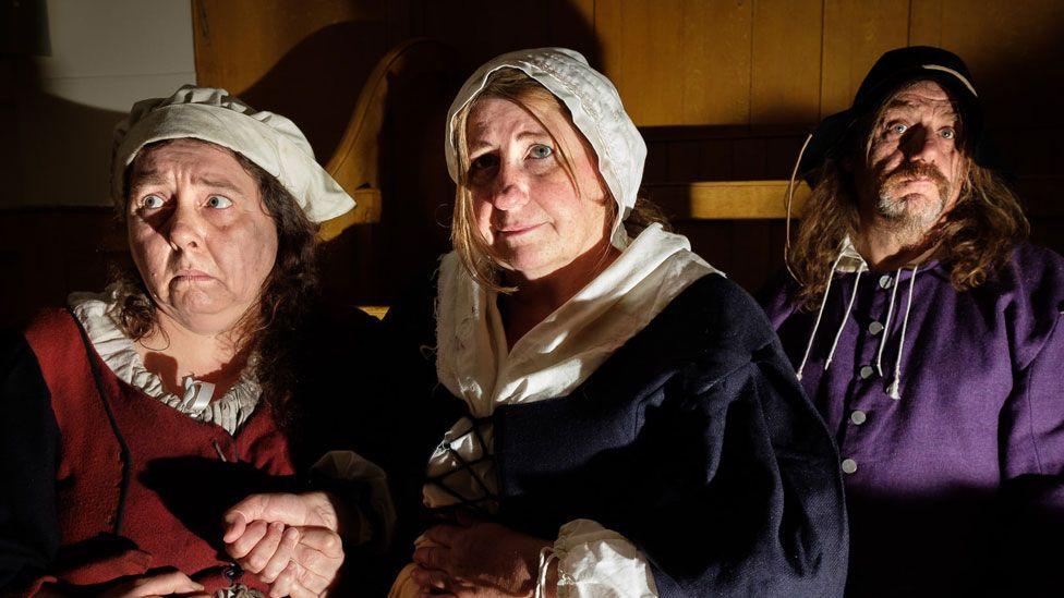 Two women and a man in mid-17th Century dress during a re-enactment of the Huntingdon witch trial. The women have white linen caps on their heads, white linen can be seen at their necks above woollen dresses, one in dark red and the other in navy. The man has a black cap on his head and a purple wool jacket. 