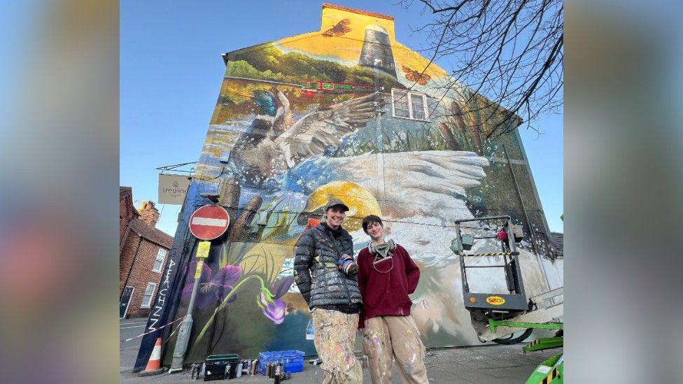 Two people wearing baggy clothes covered in paint specks smiling and standing in front of a mural they have created on the side of a dental practice. The mural depicts the River Slea with a canal boat and mill, and in the foreground is a duck and a swan splashing their wings in the water.