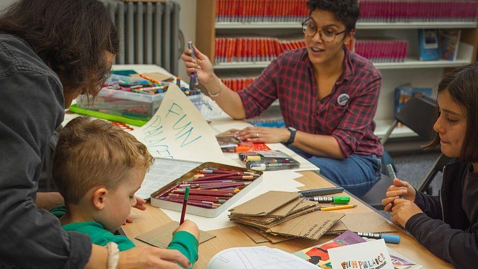 Two women sat at a crafts table with a young girl and boy. The boy is drawing on a piece of cardboard while the girl watches and there are boxes of felt tips and coloured pencils on the table