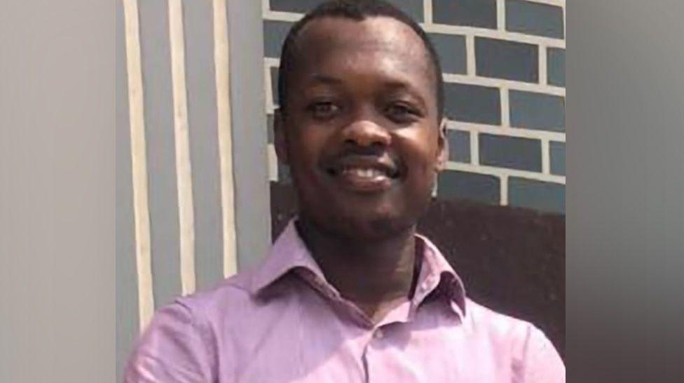 Patrice Che - a man with short black hair wearing a pink shirt and smiling. He is standing in front of a grey brick wall.