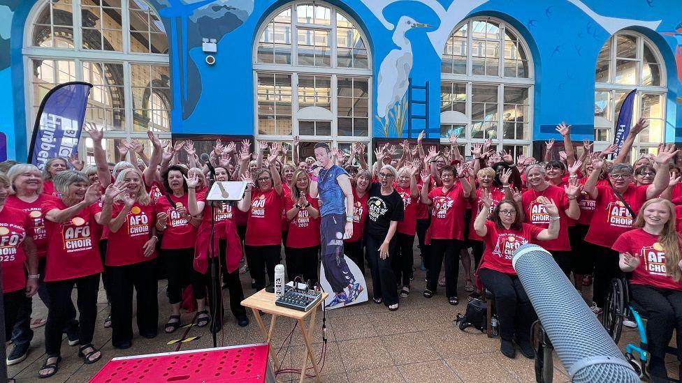 More members of the All For One Choir stood in a large group inside Paragon Station. All of them are wearing red t-shirts with the words "Disneyland Paris: AFO Choir 2025" on them in white. One woman on the front row is wearing a black t-shirt. The are posing with a cardboard cut out of Chris Martin.