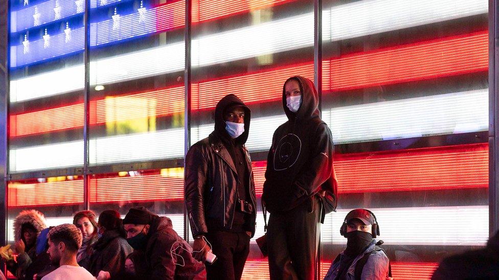 People (wearing masks and hoodies) stand in front of a large screen displaying the US flag in Times Square, New York