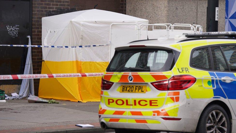 Flowers lying near two green paramedic bags and a white tent, in between two police cars.