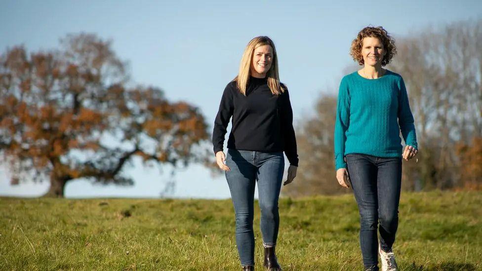 Camilla Rigby and Rachel Mostyn are walking next to each other down a grass hill. There are autumnal trees in the background. They are both wearing jeans and jumpers.