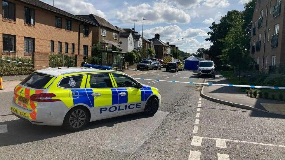 A police car sits in front of a white-and-blue tape police cordon in the middle of a road. Behind the cordon is a blue police tent and more police cars.