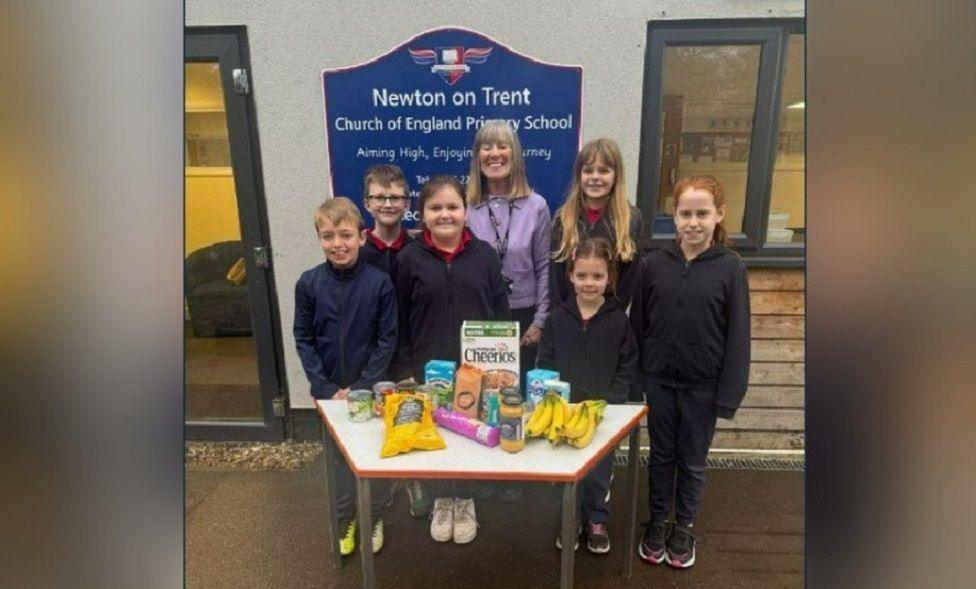 A group of four girls, two boys and a female teacher from Newton on Trent Church of England Primary School. They are standing in front of the school sign. In front of the group is a table with some food produce laid out on it including bunches of bananas and a box of Cheerios.