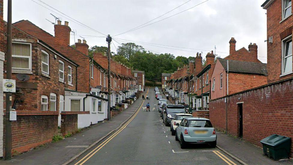 View of Vine Street in Lincoln. It is a row of red brick terrace houses along a hill with trees at the end. Cars are parked alongside the right-hand side of the road. 