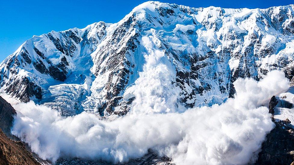 Avalanche is shown starting to form at the base of a mountain. Blue skies