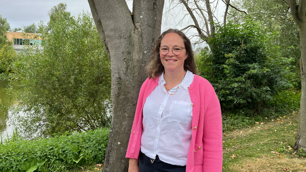 Vhari Russell pictured outside on the grass, stood in front of a tree trunk with a lake in the background. She wears thin framed circle glasses, has shoulder length brown hair and and smiles at the camera. She is wearing a white blouse and a bright pink jacket with gold buttons.