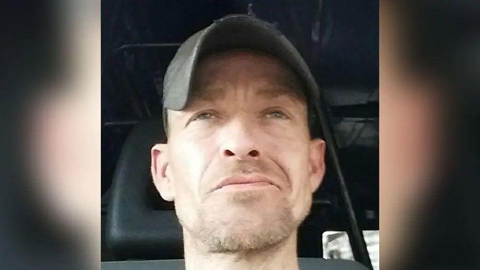 A photo of the head of a man wearing a baseball cap inside a vehicle