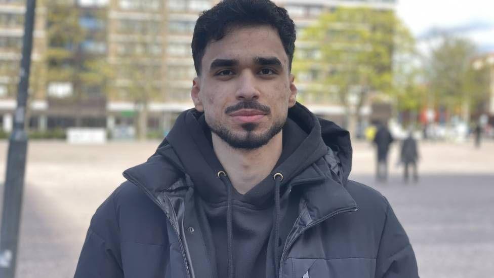 A man in a black hoodie and black coat looks at the camera. He is stood outside in a paved public area