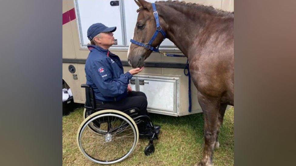 Deborah is in a wheelchair and is wearing a navy cap and a navy waterproof jacket with black trousers. She is doing a kissing face and has her hand on the chin of her horse. The horse is brown and has a white flash on his face.
