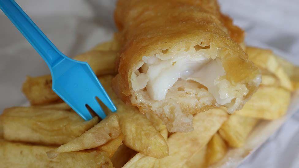 Close up of a classic portion of chip shop fish and chips. The fish is golden batter on the outside and white and flakey on the inside. A blue plastic fork is stuck in the chips