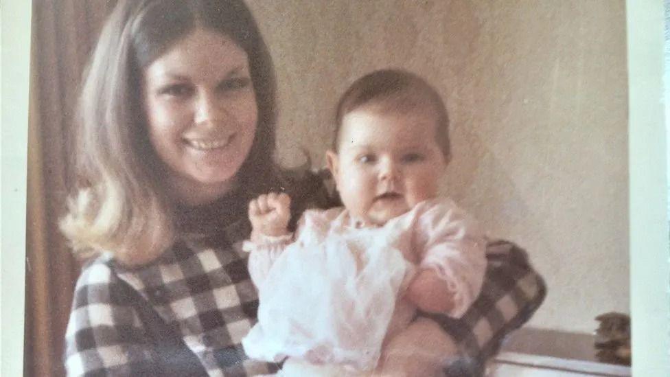 Marie is wearing a checked dress and is holding her baby in her arms.