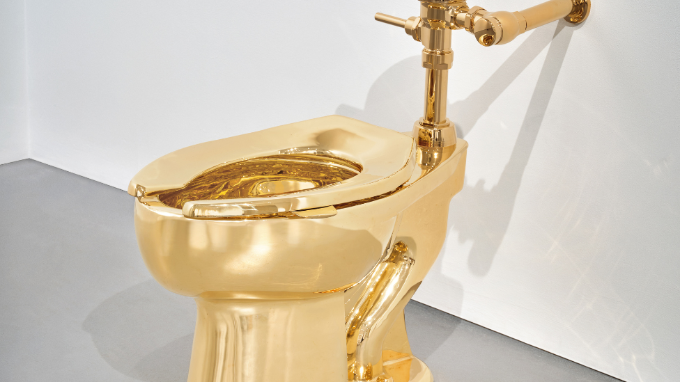 A solid gold toilet fixed on a white wall and a grey floor.