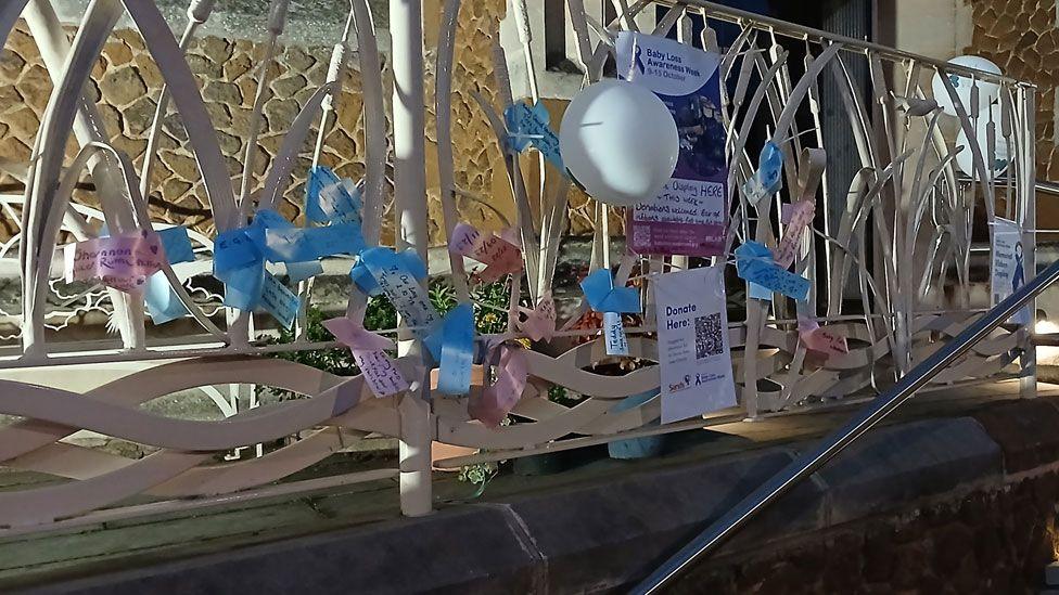 Cream railings in front of St Ives Methodist Church. Tied to the railings are blue and pink ribbons, each with writing on them. There is also a sign saying Baby Loss Week