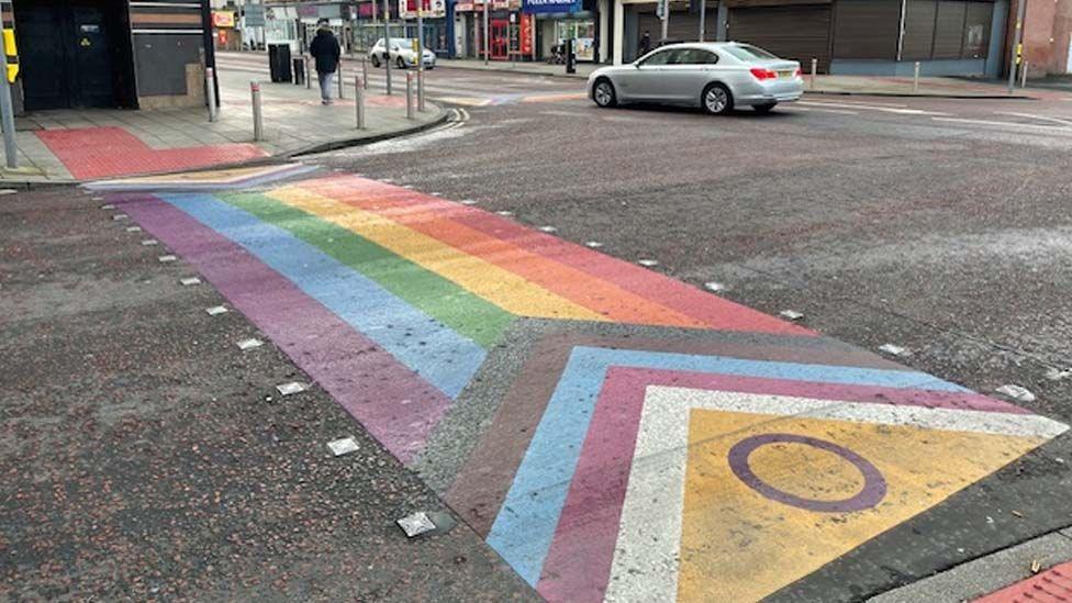 One of the rainbow zebra crossings painted on the road. A large rainbow stripe with chevrons at one end in brown, pink, blue, white and yellow