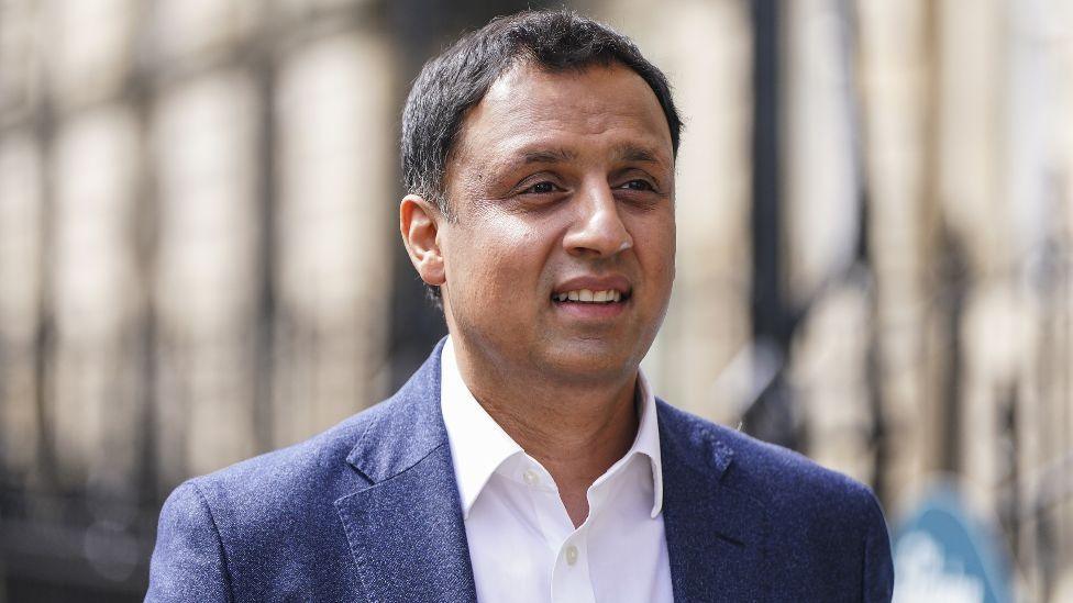 Anas Sarwar, with dark hair and wearing a blue jacket and white shite, walks along a street 