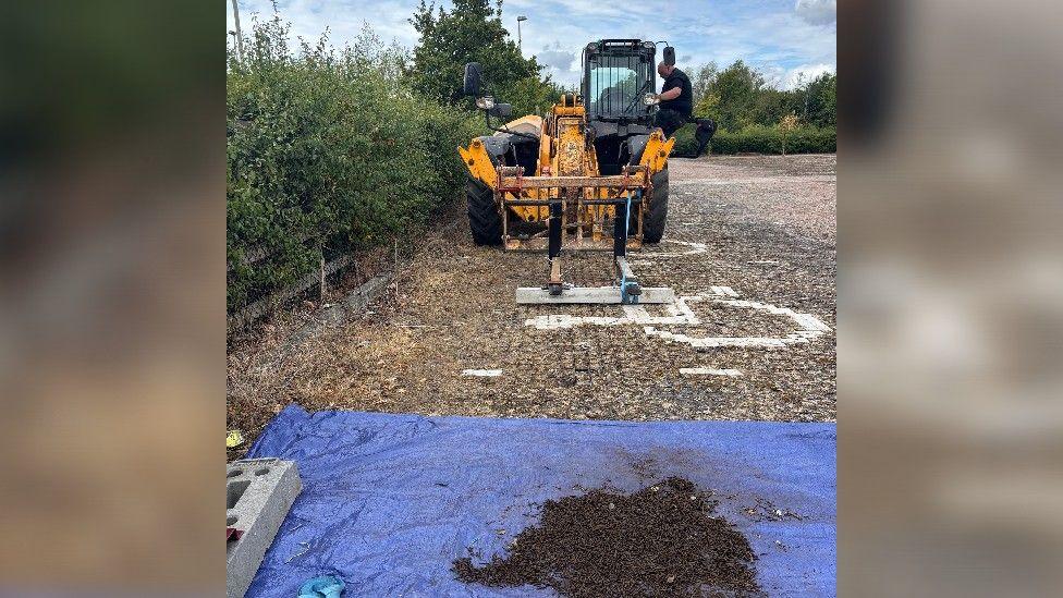 A man wearing black climbs into the cabin of a yellow digger in a disabled bay at a car boot site bordered by hedges. A metre or two in front of the digger is a piece of blue tarpaulin, weighed down by building materials at the edges, which is where recovered, rusting roofing tacks have been deposited following a clean up operation.