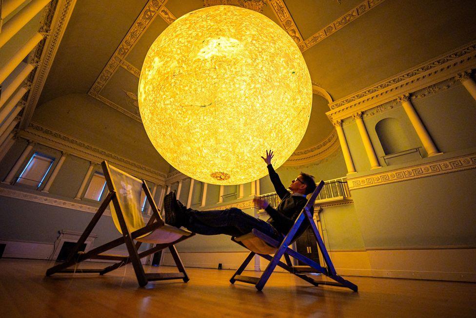 Luke Jerram sits in a chair and raises his hand towards his latest astronomical sculpture, Helios
