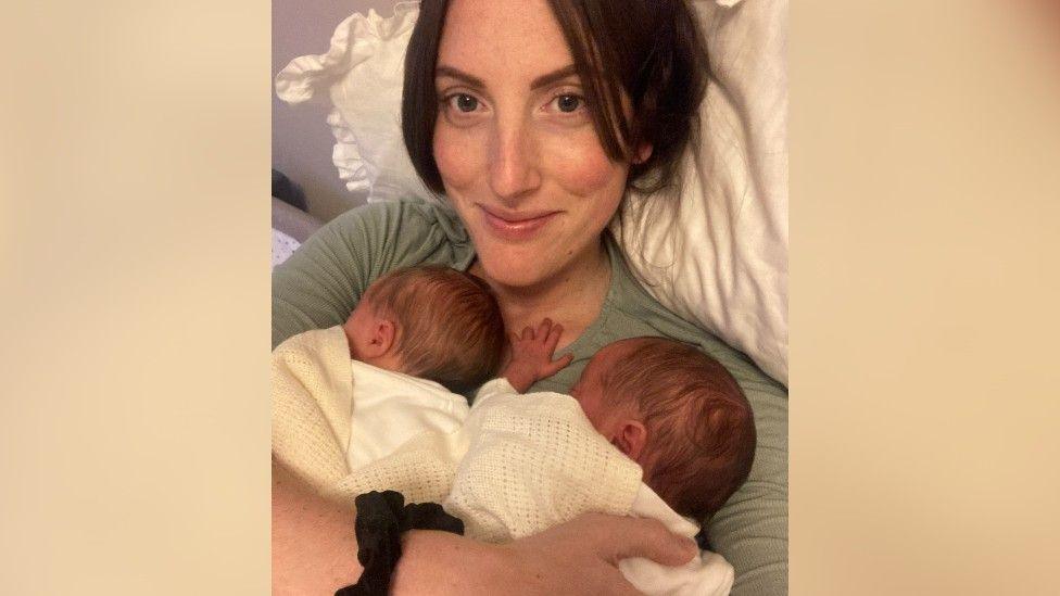 Lorna Jenkins smiles in a selfie as her newborn twin girls lay on her chest in bed. She is wearing a long-sleeved sage green top and her brown hair is loosely tied. She is propped up on a pillow and has a black scrunchie on her wrist. Her babies are wearing white, with a cream-coloured cellular blanket, with one stretching her hand out towards Lorna's neck.