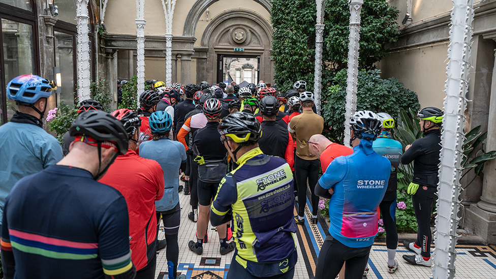 A group of cyclists in colourful kit, backs towards camera, inside Sandon Hall, listening to instructions