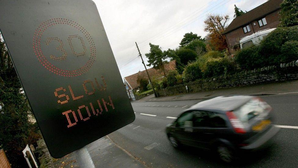 A black sign with lights reading 30 slow down at the side of a road. A slightly blurred blue car is on the road beneath the sign. A hedge in front of residential buildings on the opposite side of the street.