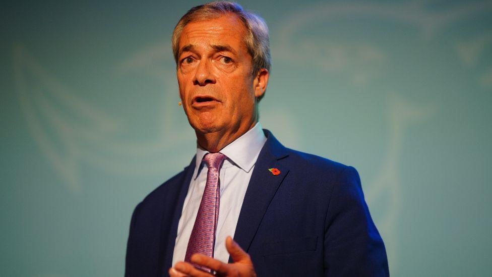 A grey-haired man with an orange complexion, wearing a dark blue suit and purple ties, stands in front of a light blue background 