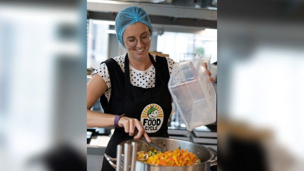 A woman wearing a blue hair net, a black and white spotty T-shirt and a black branded apron, stirring a big pot of food and smiling.