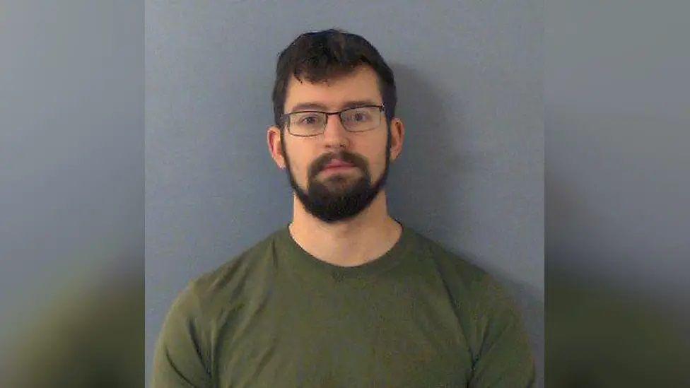 Benjamin Fields wearing a green top and glasses in a mugshot photo. He has dark hair and a thick dark beard.