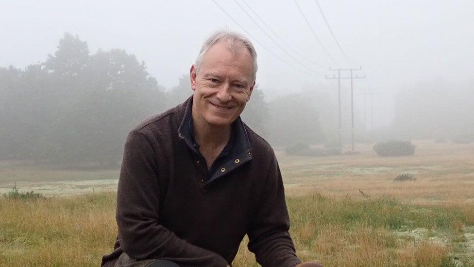 Bill Amos wearing a brown sweatshirt, smiling broadly and crouching on grassland on a misty day with telegraph poles in the distance