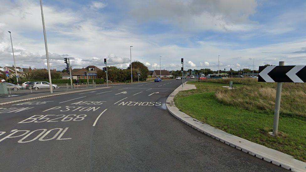 Street view image of the roundabout, with the grass verge to the right and three lanes with traffic lights to the left