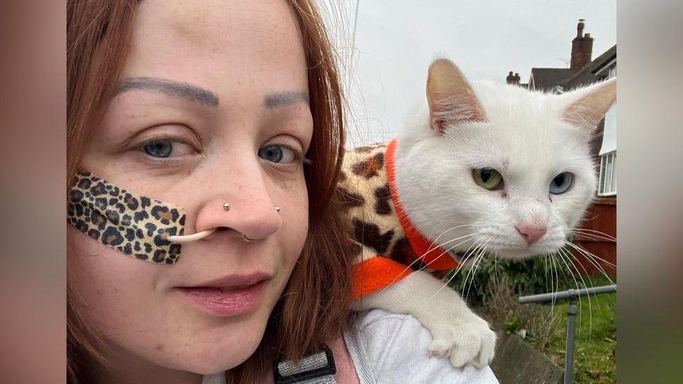 A close-up of a woman with a white cat on her shoulder. The woman has a feeding tube in her nose fastened with a leopard print plaster and red hair. The cat is wearing a leopard print pullover.