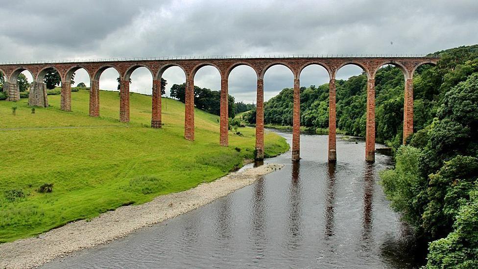 A viaduct crosses the river Tweed with one side a gently shelving field with the other a steep wooded bank