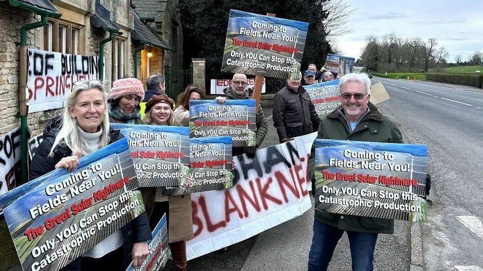 A group of about a dozen men and women standing on a pavement next to a road holding up placards opposing plans for a solar farm. Several of the placards have the words: Coming to fields near you - the great solar nightmare - only you can stop this catastrophic production. To their right is a brick building with lights in the windows.