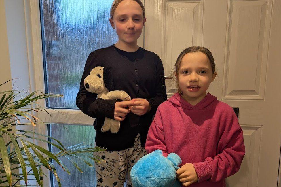 Two girls standing in front of a door inside a home they looking at the camera and are both holding soft toys.
