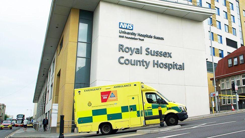 A hospital building with an ambulance in front of it. A sign on the side of the building reads "Royal Sussex County Hospital".