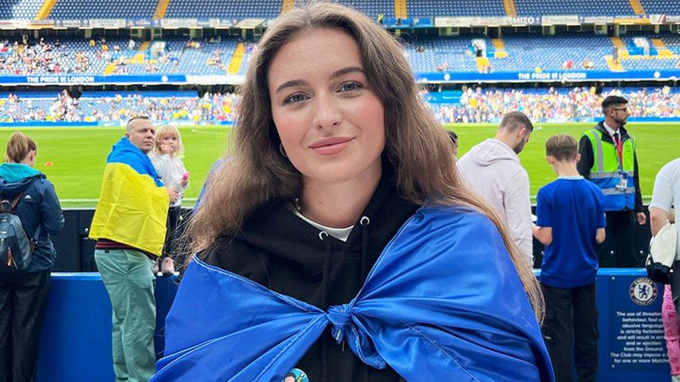 Marta, a Ukrainian woman with a blue and yellow Ukrainian flag around her looking at the camera smiling. She is wearing a black hoody. The background is of Stamford Bridge football stadium, with other people dressed in Ukrainian colours, a green grass pitch and blue seats.