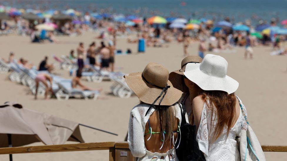 Three women with beach hats stand looking at a beach that has people sunbathing, sat under parasols, and swimming in the sea.