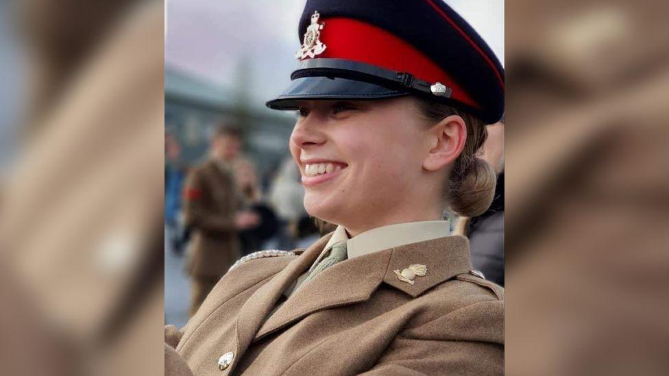 Jaysley Beck wearing a brown formal military coat and a black and red military cap. She is smiling at something off to the left of the camera.