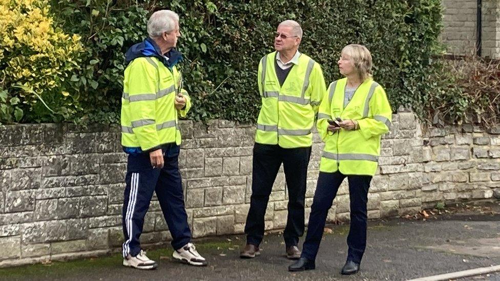 Three people in high-vis jackets stand next to a road