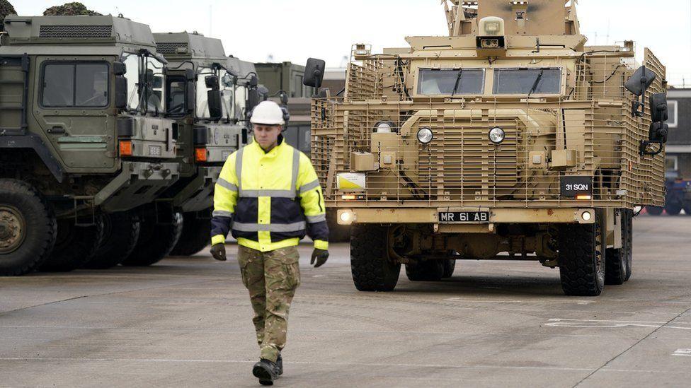 A brown coloured Mastiff armoured vehicle is being driven with a man in high-vis walking in front of it to guide it to a ship.