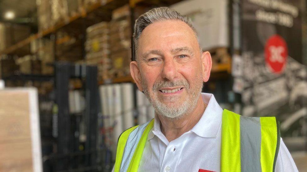 Head and shoulders of Stuart Gibbons. He has a short greying hair pulled into a slight quiff and a short white beard. He is wearing a white shirt and over it a high-vis yellow jacket. Behind him is a warehouse interior with shelves.
