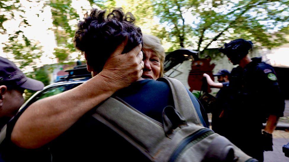 A blonde woman, who looks distressed, tightly embraces a man with short brown hair. Her hand is round the back of his head and only the back of his head, back and rucksack can be seen.