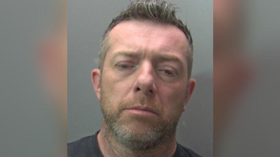 Scott Walker is seen in a police custody shot. He has short, light brown hair and a stubbly beard and moustache. He is wearing a dark T-shirt