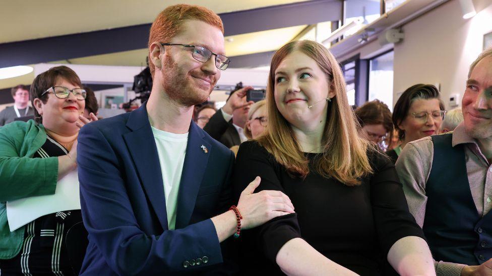 Ross Greer, who has ginger hair and glasses, and Gillian Mackay, who has fair hair and is wearing a black jumper, sit next to each other in the front row of an audience. Greer, wearing a navy suit jacket over a white t-shirt, has his right hand on Mackay's upper right arm in a friendly gesture. They are smiling towards each other, with audience members clapping in the background.