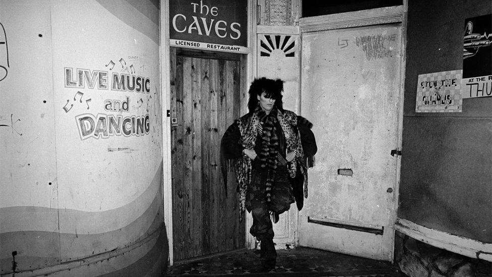 Black and white photograph of Peter Burns stood between two doors under as sign reading The Caves and painted writing on the wall which says Live Music and Dancing