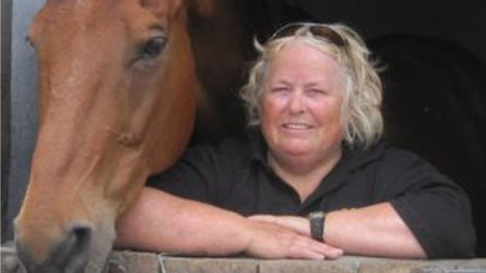 Wendy Buckney leans on a stable door, with a brown horse next to her. Wendy has blonde, shoulder length hair and is smiling at the camera. She is wearing a black T-shirt