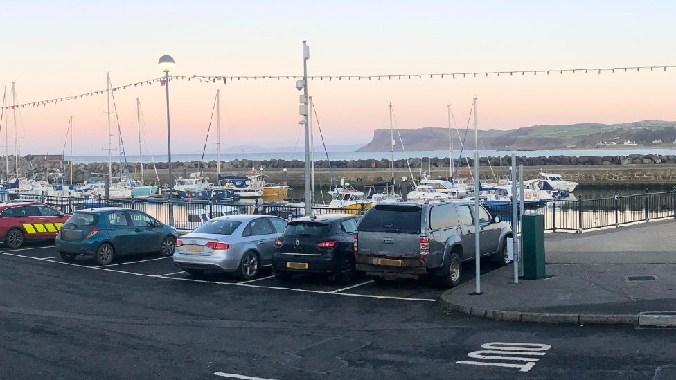 Car part in Ballycastle with a number of cars parked alongside marina, boats, water and mountains are visible in the background, the sky is at sunset and there are string lights attached on poles around the car park